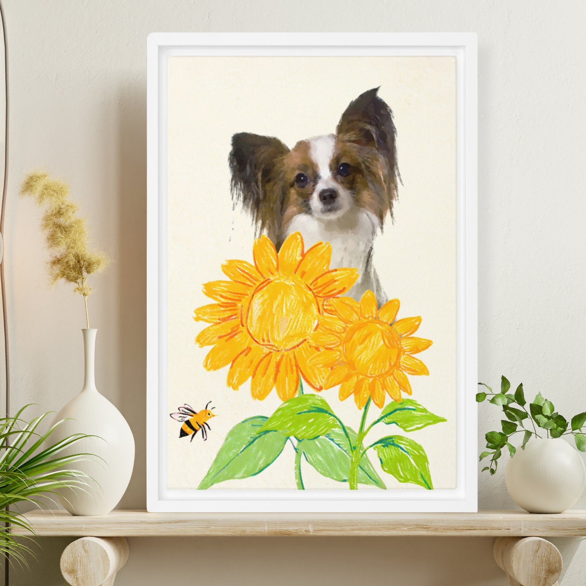 Minimalist crayon-style pet portrait of a Papillon dog with soft watercolor features, surrounded by bright yellow sunflowers and a bee, displayed in a light wooden frame next to a green potted fern.