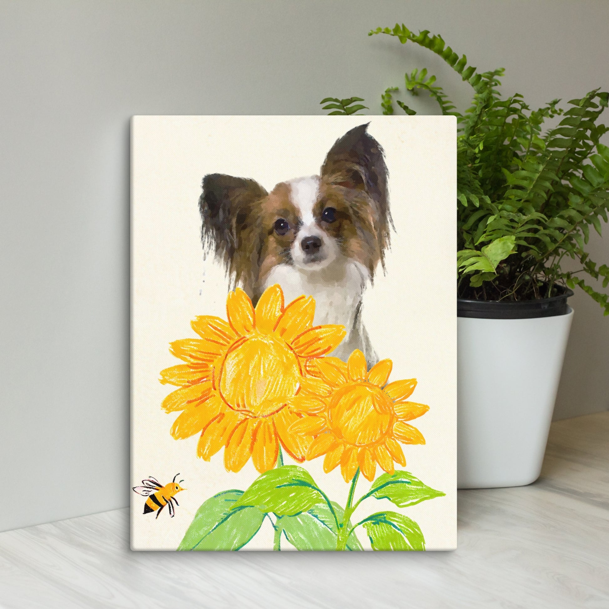 Minimalist pet portrait of a Papillon dog in soft watercolor style, surrounded by bright yellow crayon-style sunflowers and a bee, set against a light canvas and displayed beside a green potted fern