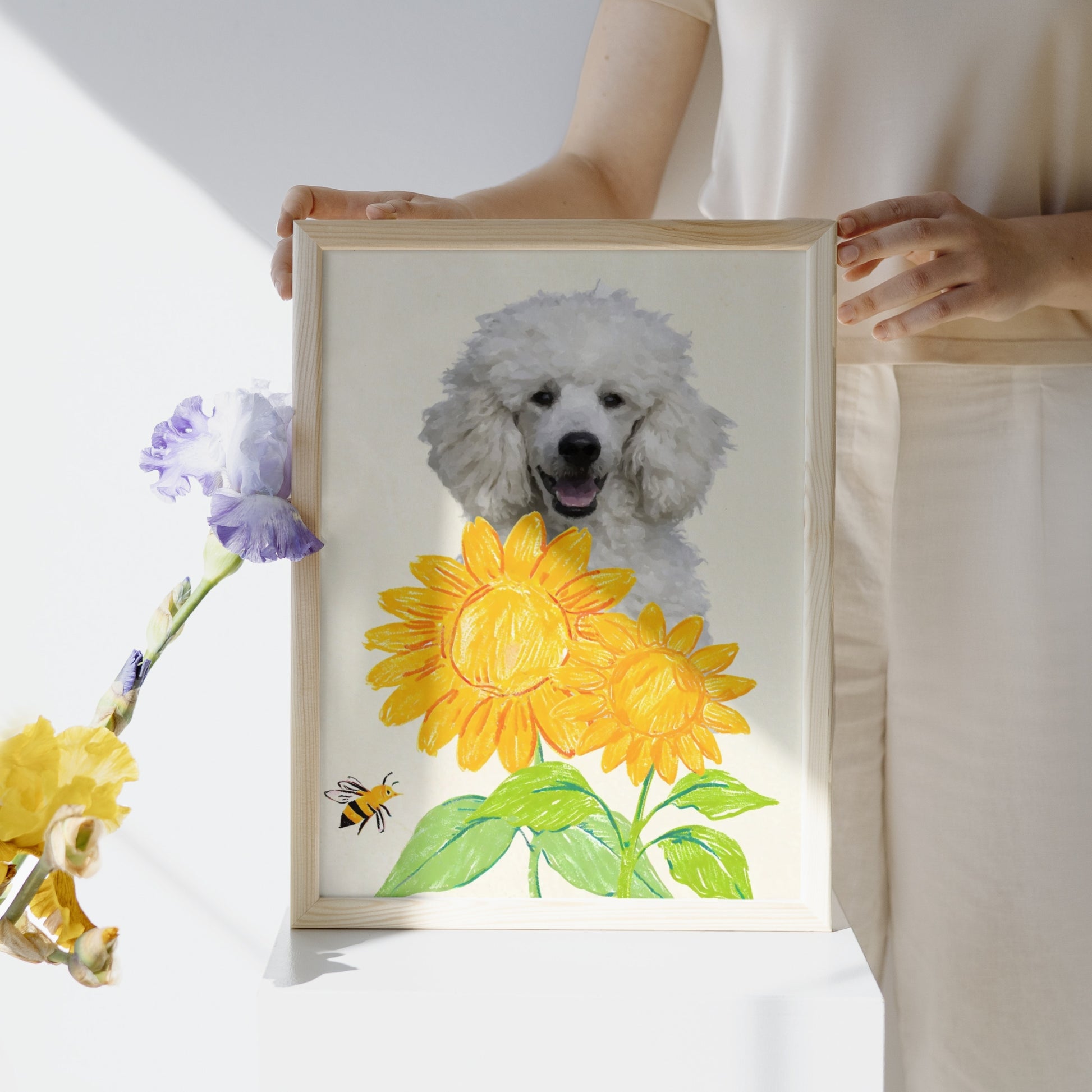 Minimalist pet portrait of a smiling white poodle in soft watercolor style, framed by bright crayon-style sunflowers and a bee, with a purple and yellow flower to the side, displayed in a light wooden frame held by a person.