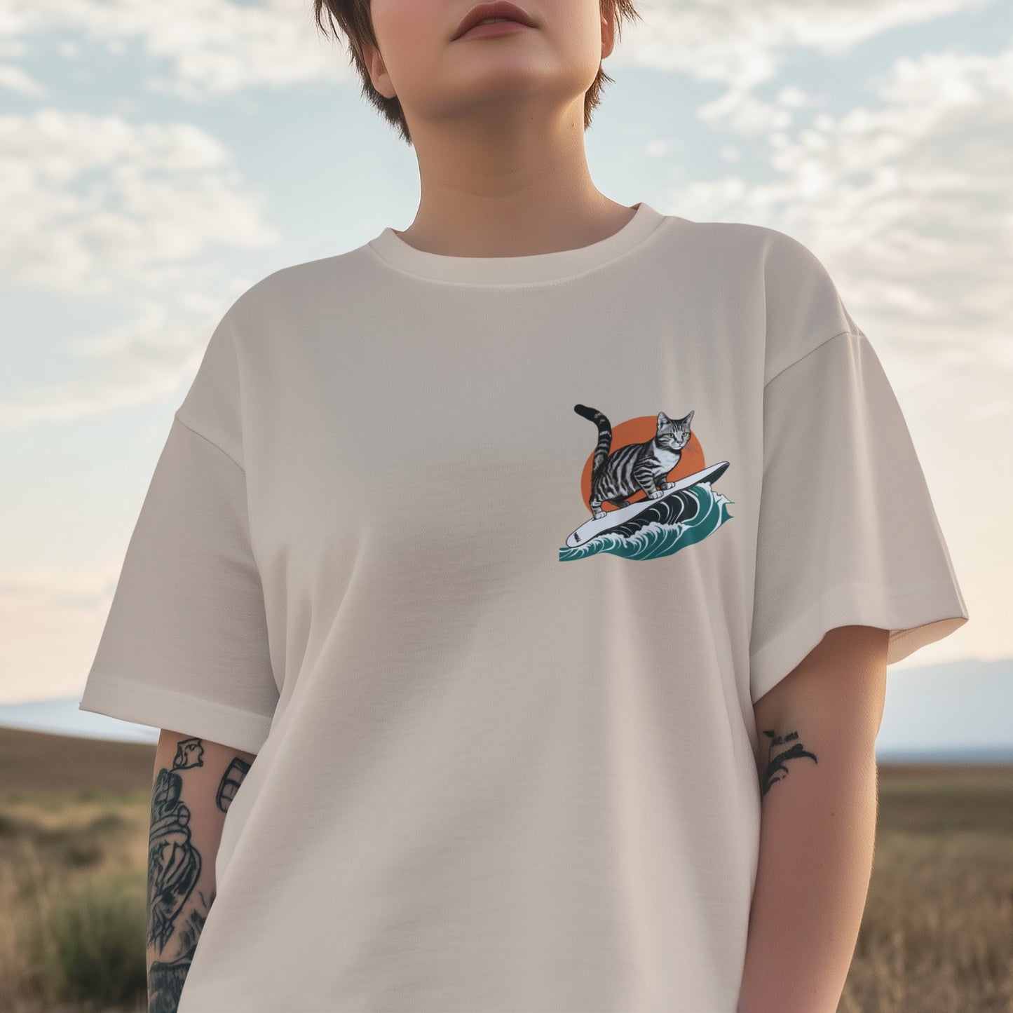 A young woman wearing a beige colored cotton minimalist graphic t-shirt featuring a small surfing cat graphic on the front left chest.