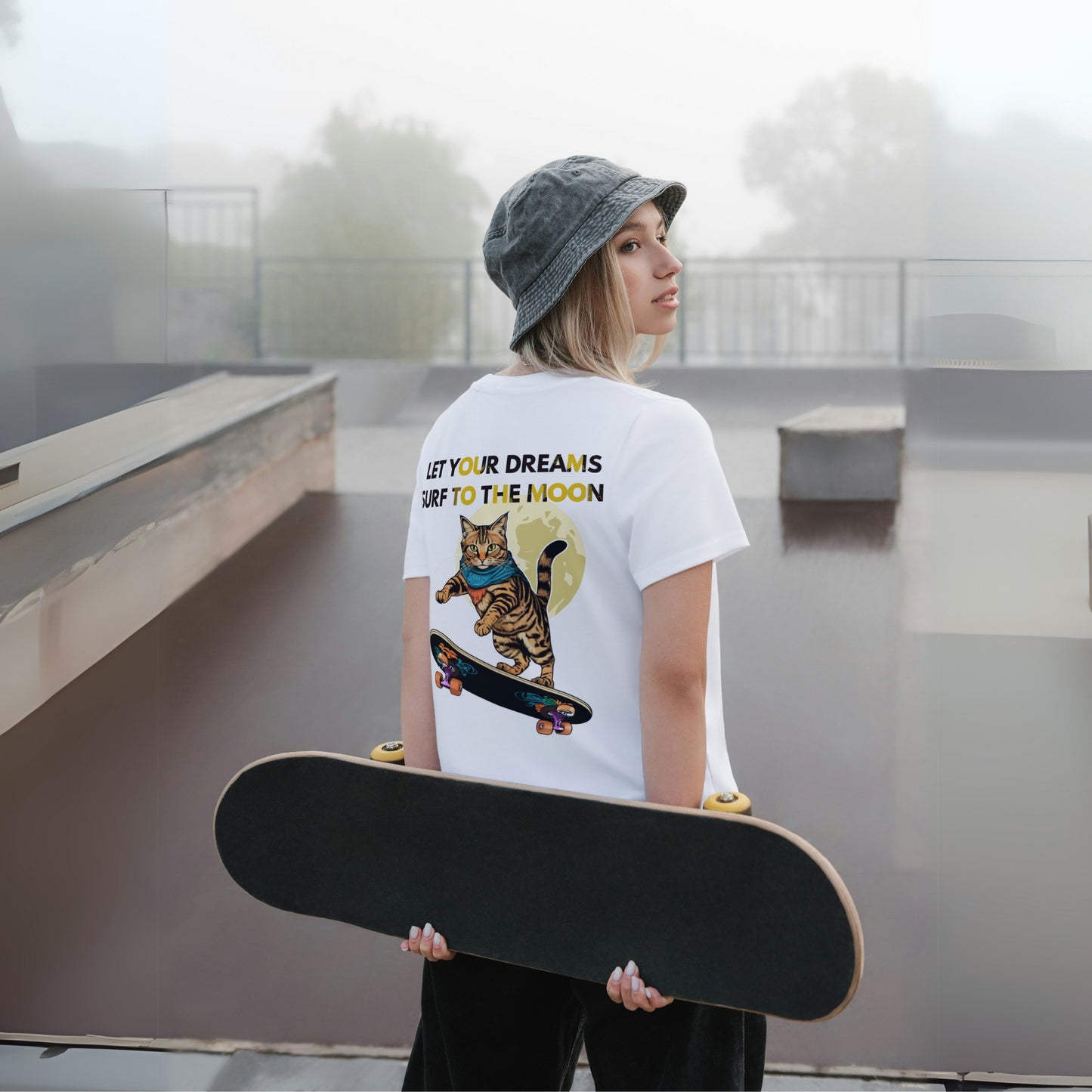 Back view of a young girl wearing a white cotton T-shirt featuring a cat skateboarding toward the moon. She holds a skateboard while posing for a photo, showcasing the fun, dynamic design.
