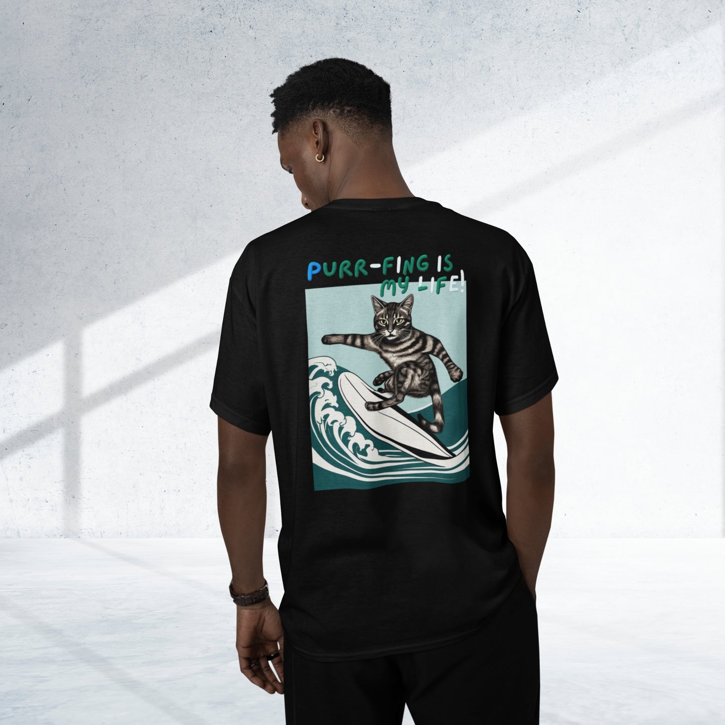 From the back view, a young man sports a stylish black cotton short-sleeve t-shirt adorned with a whimsical design of a surfing cat and the amusing phrase 'Purr-fing is my life!' on the back, capturing his joy and humor.