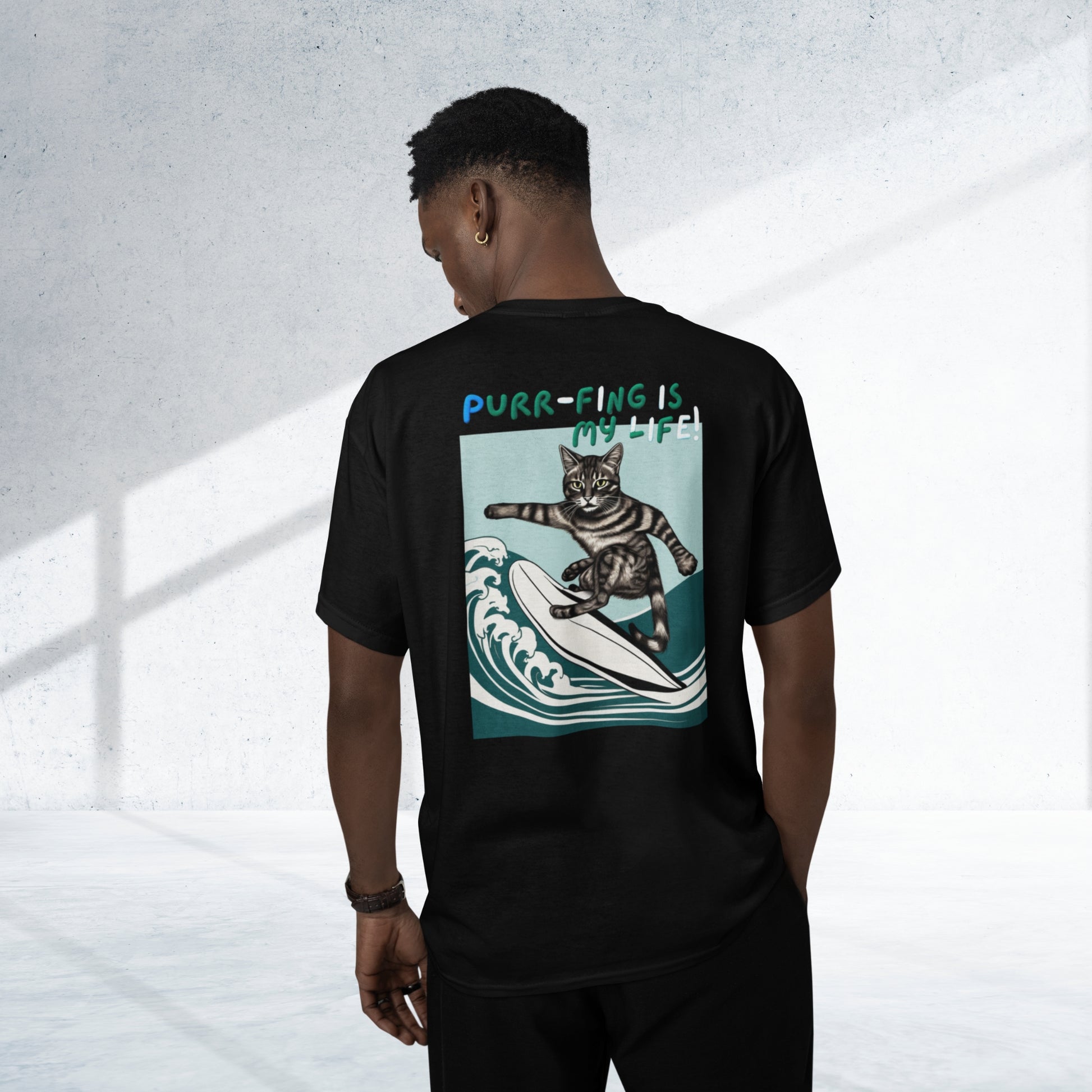 From the back view, a young man sports a stylish black cotton short-sleeve t-shirt adorned with a whimsical design of a surfing cat and the amusing phrase 'Purr-fing is my life!' on the back, capturing his joy and humor.
